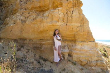 Makyaj yapan güzel bir genç kadın, bej bir elbise ve deniz kenarındaki bir uçurum duvarına dayanmış altın mücevherler. Kadın farklı pozlar ve ifadeler veriyor. Güzellik ve moda kavramı