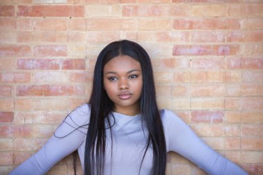 Afrikalı bir kadın, genç ve güzel, bej bluzlu ve siyah deri şortlu. Kolları açık, kırmızı tuğladan bir duvara yaslanmış.