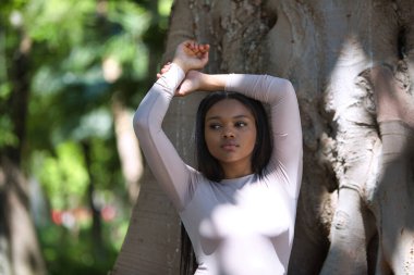 Uzun kahverengi saçlı, genç ve güzel, sıradan kıyafetler giymiş Afrikalı bir kadın kollarını kaldırıyor ve güneş ile gölge arasındaki incir ağacının gövdesine yaslanıyor.