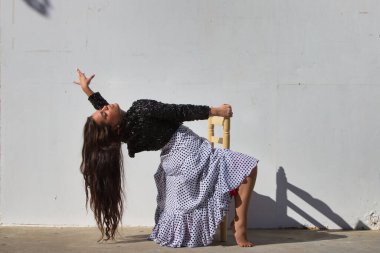 Latin kadın, genç ve güzel, dans eden flamenko, eski tahta bir sandalyede oturuyor ve kafasını geriye atıyor. Kadın, siyah benekli ve siyah ceketli, beyaz, dalgalı bir elbise giyiyor.