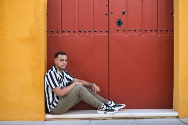 Çizgili gömlek ve kargo pantolonu giyen sakallı, genç, yakışıklı bir adam Seville, İspanya 'da kırmızı kapısı olan bir evin merdivenlerinde dinleniyor. Adam turist olarak geliyor.
