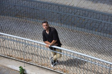 Sakallı, siyah tişörtlü ve bej kargo pantolonlu genç, yakışıklı bir adam, metal bir korkuluğa tutunuyor, rampadan aşağı indi ve yukarı bakıyor. Fotoğraf yukarıdan çekildi