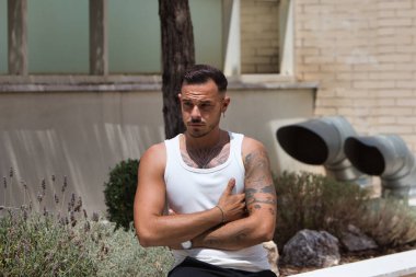 Sakallı, dövmeli, küpeli, beyaz atletli, siyah pantolonlu, çiçek tarhında oturan, kolları çaprazlanmış, ciddi görünen genç bir adam.