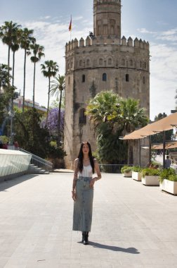 Kollarında dövmeleri olan, sıradan kıyafetler giyen, güneşli bir günde şehrin büyük bir caddesinde poz veren genç ve güzel bir Latin kadın. Arka planda Sevilla 'nın altın kulesi var.