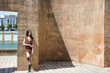 Latin bir kadın, genç ve güzel esmer, kollarında ve bacaklarında dövmeleri olan, kahverengi bir elbise ve siyah çizmeler giyen, kahverengi beton bir duvara yaslanan. Kadın rahatladı.