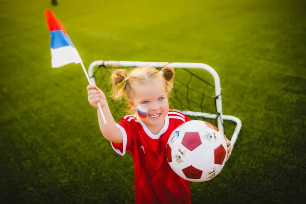 Little funny blonde girl holding huge russian flag and ball scream, standing in front of football gate. High quality photo