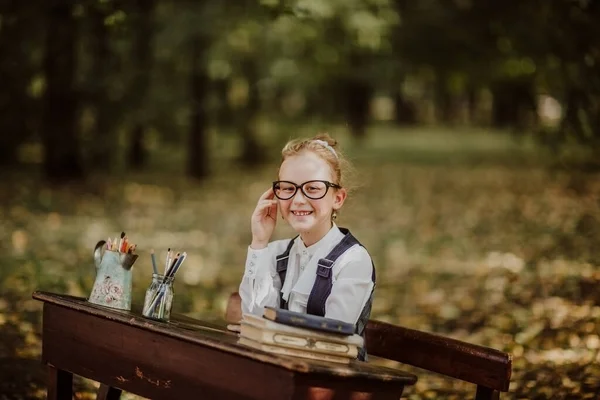 Portrait of young funny school girl in glasses sitting on a wooden desk outside. Back to school. Copy space