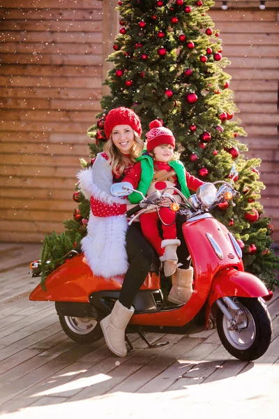 Beautiful blonde mom and son in Christmas red sweaters and red hats sit on a red vintage moped on the background of a Christmas tree under the flying snow. High quality photo