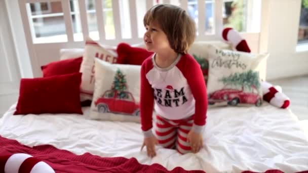 Cute boy in Christmas pajama sits on a white bed with Christmas pillows. It's snowing outside. Slow motion — Stock Footage Cute Boy Christmas Pajama Sits White Bed Christmas Pillows Snowing Royalty Free Stock Video