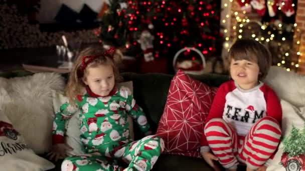 Cute boy and girl in Christmas pajama sitting on the sofa with Christmas pillows on the background of Christmas tree and fireplace. A boy kisses a girl. — Stock Footage Cute Boy Girl Christmas Pajama Sitting Sofa Christmas Pillows Background Stock Video