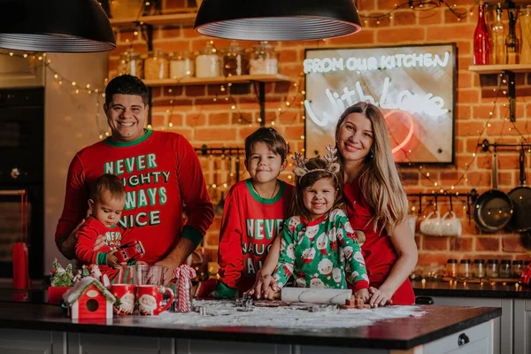 Young family of five in Christmas pyjamas making cookies on the kitchen with Christmas background.