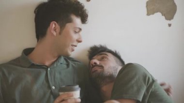 Eşcinsel erkek çift yatakta uzanıyor ve birbirlerine sarılıyorlar. Çocuk kahve içiyor. LGBT