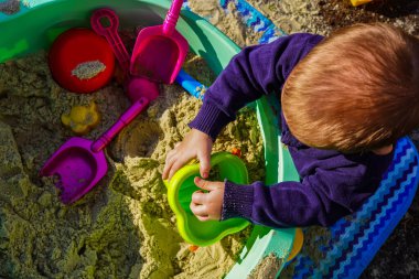 Çocuk kum havuzunda oynuyor. Kürekler, kalıplar, çocuk oyuncakları. Küçük kızlar ve oğlanlar için bir oyun. Çocukluk, eğlence ve tasasız zaman geçirmek için harika bir zamandır. Kum