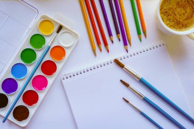 Çizim üzerinde çalışmak için bir yer. Suluboya resimler, albüm, fırçalar, renkli kalemler. Aromatik kahveli kupa. Beyaz ışık arka planı. Çizim eğitimi. Yaratıcılık için sanat malzemeleri.