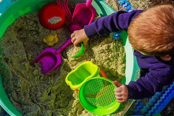 Çocuk kum havuzunda oynuyor. Kürekler, kalıplar, çocuk oyuncakları. Küçük kızlar ve oğlanlar için bir oyun. Çocukluk, eğlence ve tasasız zaman geçirmek için harika bir zamandır. Kum