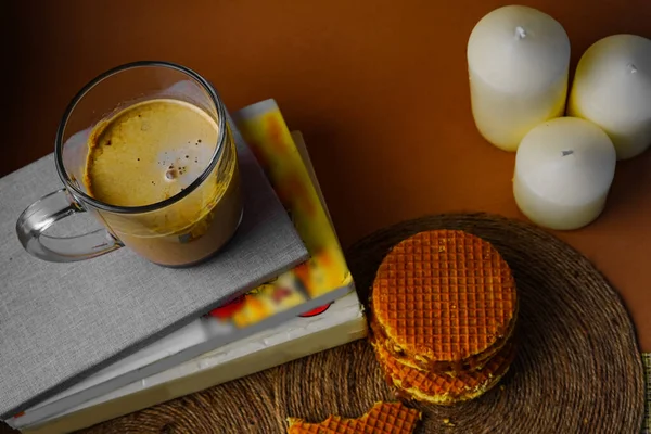 Kahverengi arka planda kremalı kahve fincanı. Sıcak bir içeceğin güzel bir resmi. Lezzetli fotoğraf. Yoğunlaştırılmış sütlü tatlı yuvarlak waffle 'lar. Alışılmadık atıştırmalıklar, kahvaltı, öğle yemeği. Tatlı. Bir yığın kitap. Üç mum..