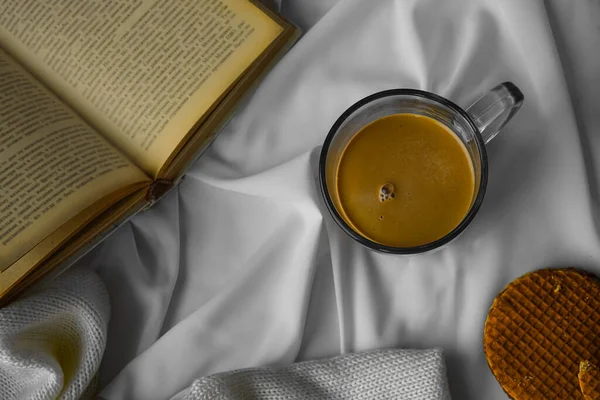 Beyaz çarşafta kremalı bir fincan kahve, hafif örülü bir battaniye. Sıcak ve aromatik bir içecekle günaydın. Kitap gri bir kapakta. Fotoğraf yukarıdan çekildi. Yuvarlak waffle 'lar yoğunlaştırılmış sütle evlenir. Düz Kopyalama alanı