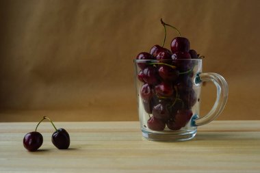 Hafif doğal ahşaptan yapılmış bir masanın üzerinde taze kirazlarla dolu şeffaf bir cam bardak var. Yakınlarda iki kiraz böğürtleni var. Lezzetli meyveler sağlık ve diyet için iyidir..