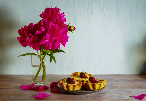Açık ahşap bir masanın üzerinde su dolu cam bir vazo, üç parlak pembe şakayık çiçeği ve kirazlı kekler var. Yaprakların etrafından. Güzel yemek arkaplanı resmi