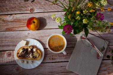 Lezzetli bir öğle yemeği, güzel bir ahşap masada sunulur. Şirin bir buket kır çiçeği, olgun bir şeftali, aromatik bir kahve ve çikolatalı fıstık ezmeli bir sandviç, gri bir kitap, bir defter ve bardaklar.