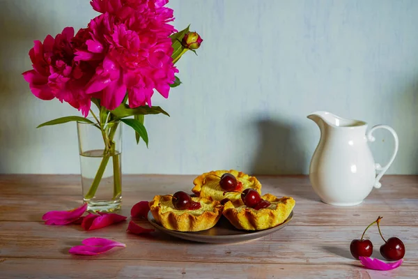 Açık ahşap bir masanın üzerinde su dolu cam bir vazo, üç parlak pembe şakayık çiçeği, kirazlı kekler ve bir sürahi süt var. Yaprakların etrafından. Güzel yemek arkaplanı resmi