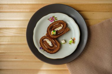 Fotoğraf en üst açıdan çekilmiş. Doğal ahşap tahtalardan yapılmış hafif bir masada bir pamuk peçete ve lezzetli ev yapımı hamur işleriyle dolu bir tabak var. Yuvarlak şeklinde bir pasta.
