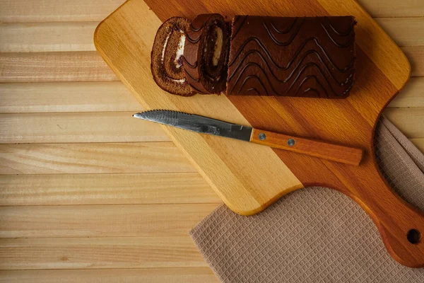 Doğal ahşap tahtalardan yapılmış hafif bir masada bir pamuk peçete ve üzerinde bir bıçağın bulunduğu bir tahta kesme tahtası var. Yuvarlak ve ev yapımı taze hamur işleri şeklinde lezzetli bir çikolatalı kek.