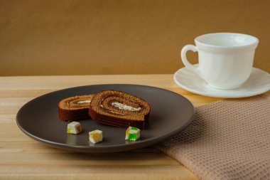 Gri bir tabakta, yanında bir fincan sıcak aromatik kahve bulunan rulo şeklinde iki dilim lezzetli çikolatalı kek ve üç küçük marmelat var. Hafif ahşap bir masada servis edilen tatlı.