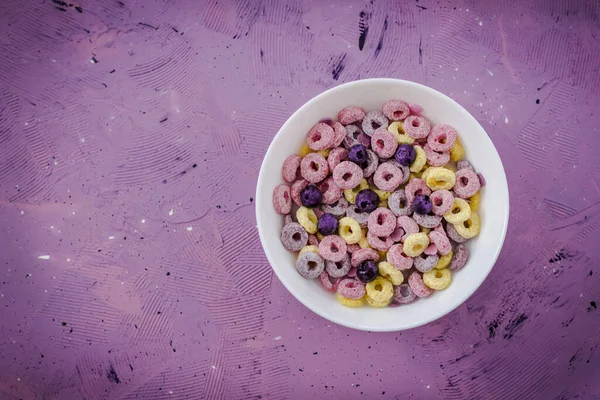 As if in space! Healthy cereal loops with blueberry in a bowl. Multicolored loops on decorative violet background. Quick breakfast or snack, flat lay.