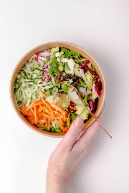 Kraft kağıt paketinde, beyaz masada, kadın eli ile sağlıklı yassı salata. Şeritler halinde kesilmiş sebzeler ve otlar. Spor yemeği. - Öğle yemeği. Beslenme kavramı