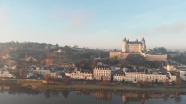 Saumur kasabası, ortaçağ şatosu ve kilisesiyle gündoğumunda nehre yansıyan, Loire Vadisi, Fransa. Hava aracı atışı, sola dönüyor ve nehrin üzerinde güneşin doğuşunu gösteriyor.