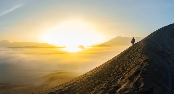 Man with obscured face walking on the edge of a crater slope , over a sea of clouds at sunrise, in Bromo volcano, Java, Indonesia. The double crater is a popular touristic destination for a sunrise trek.
