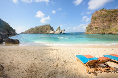 Popüler fotoğraf mekanı ve turizm merkezi Atuh Sahili. Tropikal bir kumsalda boş şezlonglar, uzakta deniz taşları ve uçurumlar, ve Nusa Penida, Bali, Endonezya 'da turkuaz deniz.