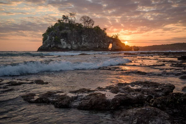Kristal Koyu 'nda gelgitle birlikte gün batımı kaya adası boyunca ilerliyor. Deniz suyu Nusa Penida, Bali, Endonezya' daki kaya ve su birikintilerinin üzerine dökülüyor.