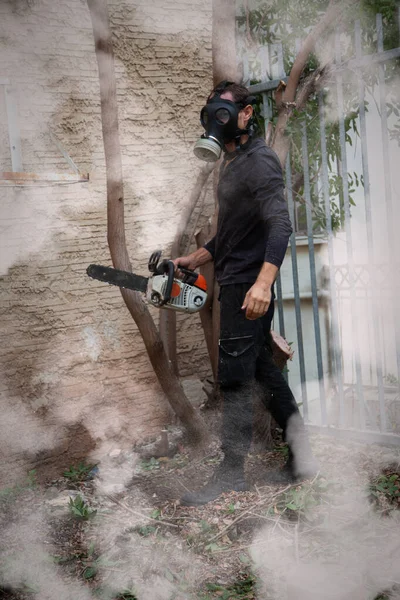 Man in a gas mask in the middle of the chainsaw\'s smoke. Dangerous job. Powerful chainsaw. Lumberjack hold chainsaw. Gardener lumberjack equipment. Working in the garden near home during quarantine.
