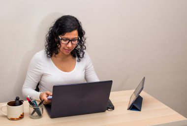 Beyaz tişörtlü, gözlüklü, siyah saçlı genç bir kadın cep telefonundan mesaj gönderirken bilgisayarını ve tabletini masaya bırakıp evden çalışıyor..