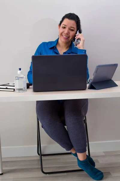 Beyaz kadın, mavi resmi gömlek ve pijama pantolonuyla evden çalışıyor. Laptopunda uyku çorapları, masada tablet ve sağında iki siyah defter ve telefonda konuşurken el dezenfektanı var..
