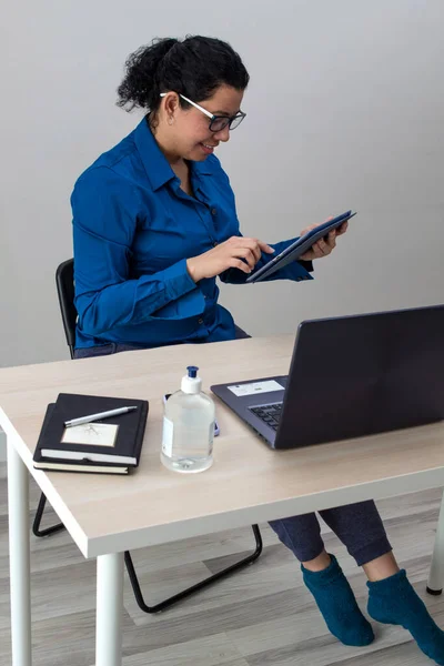 Caucasian woman working from home in a blue formal shirt and pajama pants with sleeping stockings on her laptop and tablet on a table and to her right two black notebooks and a hand sanitizer while talking on the phone.