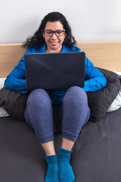 Young caucasian woman working from home on her bed in a blue formal shirt and night pants and stockings while on a work conference