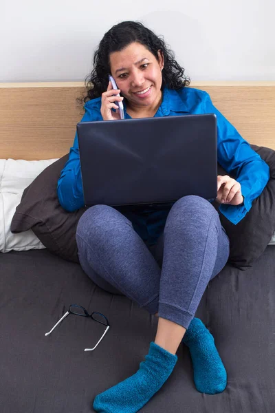 Young caucasian woman working from home on her bed in a blue formal shirt and night pants and stockings while talking on her mobile work phone