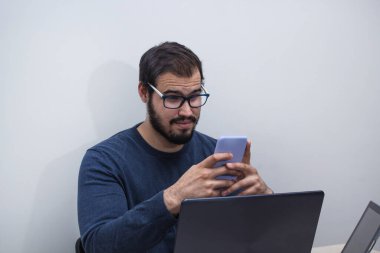 Gözlüklü ve mavi kazaklı genç beyaz adam telefonda iş arkadaşıyla konuşurken evden çalışıyor..