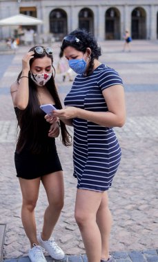 Şehirdeki bir meydanda elektronik cihazlarını kullanırken tıbbi maske takan genç kadınların seçici odak noktası