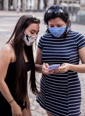 Şehirdeki bir meydanda elektronik aletlerini kullanarak tıbbi maske takan genç kadınların seçici odak noktası