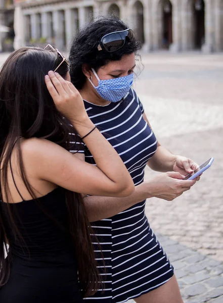 Şehirdeki bir meydanda elektronik aletlerini kullanarak tıbbi maske takan genç kadınların seçici odak noktası