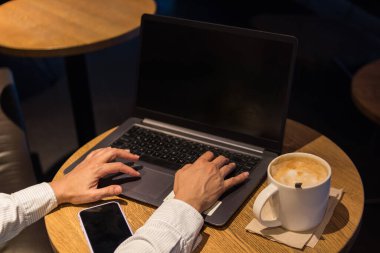 Bir kahve dükkanında, ahşap bir masada dizüstü bilgisayar, yanında bir fincan kahve ve yanında telefon.