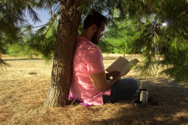 Kot pantolonlu ve pembe gömlekli beyaz bir adam ormanda bir ağacın yanında oturmuş siyah bir kitap okuyor.