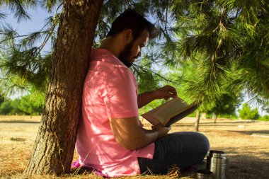 Kot pantolonlu ve pembe gömlekli beyaz bir adam ormanda bir ağacın yanında oturmuş siyah bir kitap okuyor.