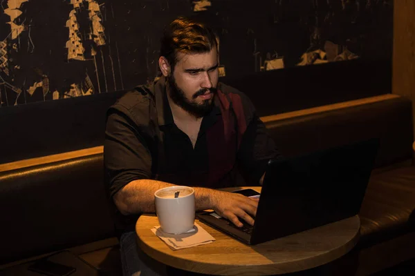 Siyah gömlekli, laptopuyla çalışan, kahve içen ve telefonuyla dolaşan beyaz bir genç.
