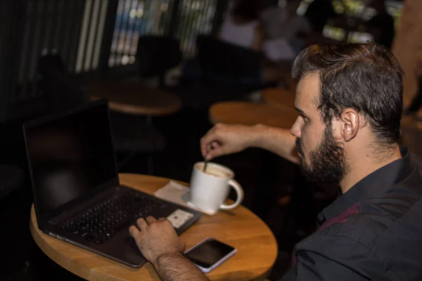 Siyah gömlekli, laptopuyla çalışan, kahve içen ve telefonuyla dolaşan beyaz bir genç.