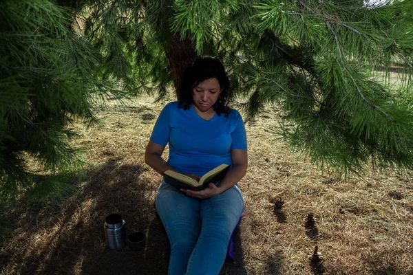 Kot pantolon giyen, mavi kazaklı beyaz bir kadın yanında termosla tarlada siyah bir kitap okuyor.